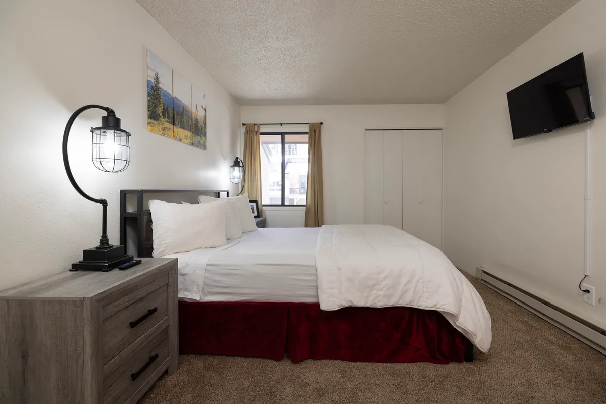 A cozy bedroom interior with a plush bed, soft linens, a nightstand with a lamp, a window offering natural light, and stylish artwork above the headboard. The scene exudes a warm, calming ambiance suitable for unwinding after a busy day.