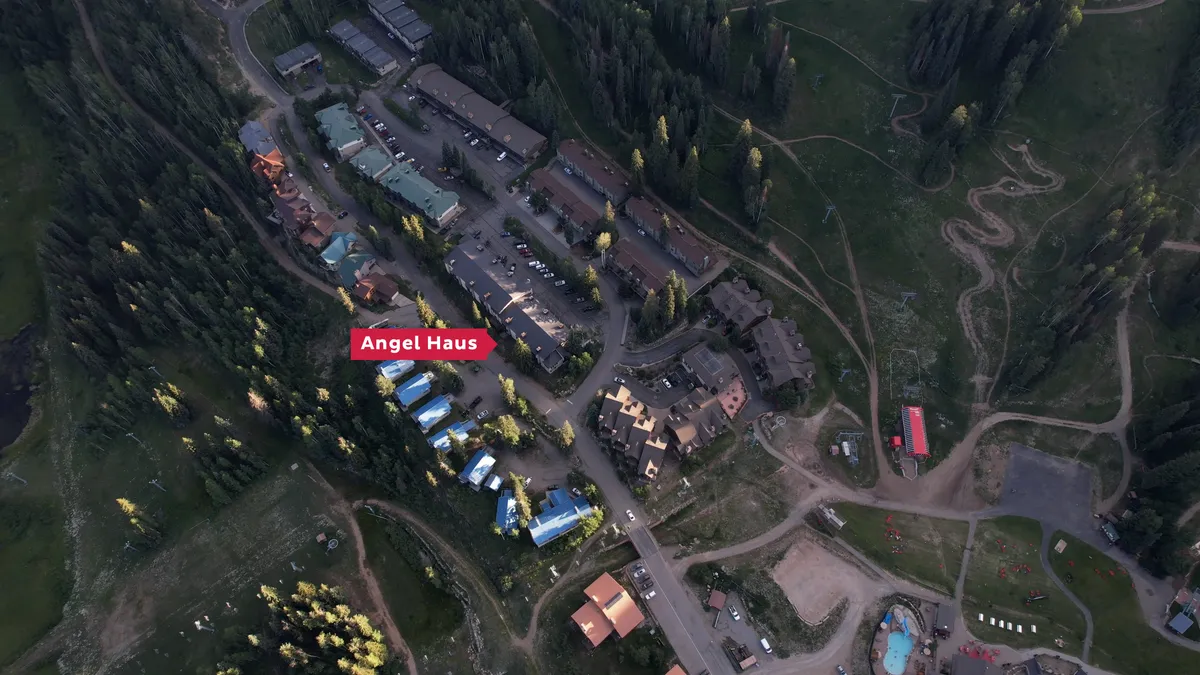 A daylight exterior view showcasing the Angel Haus building and surrounding lodges, with winding paths leading through forested areas. The environment is inviting and hints at outdoor adventures and moments of peaceful reflection.