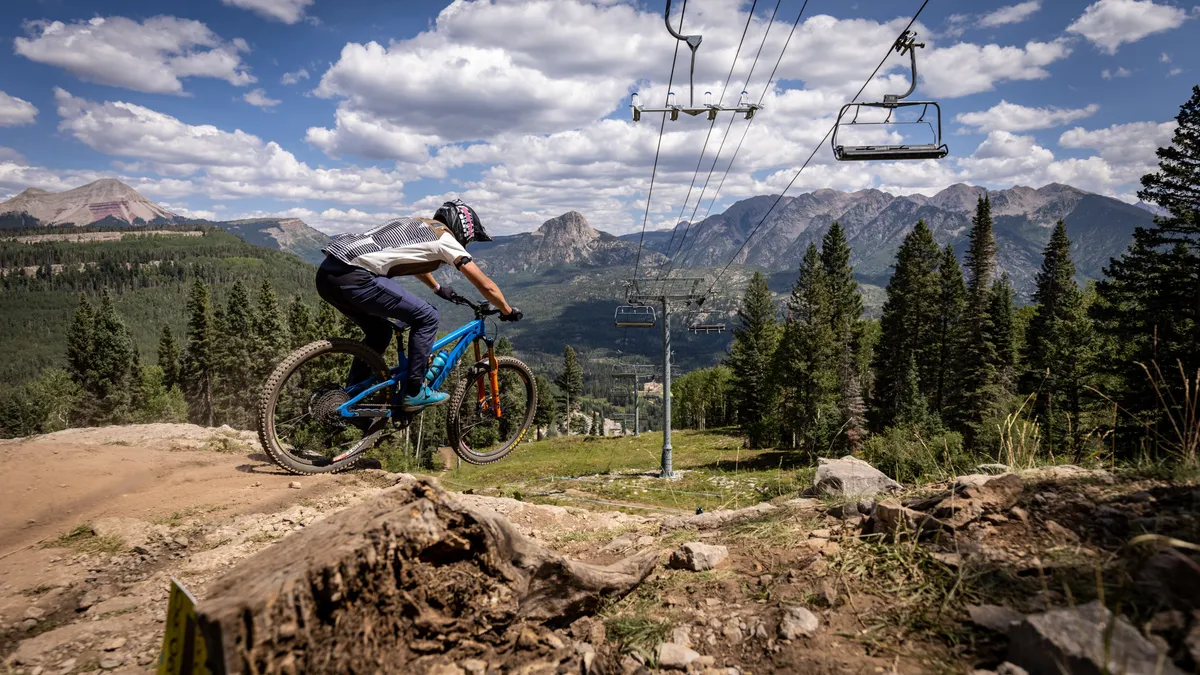 A mountain biker rides along a forested mountain trail with a breathtaking mountain range and a ski lift visible in the background. Lush trees and a dense forest envelop the area, highlighting multiple outdoor activities possible in all seasons.