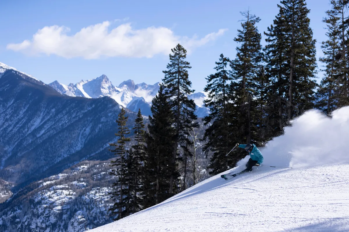 A skier carves down a snow-covered slope flanked by tall pine trees, with breathtaking mountains rising in the background. The action is upbeat and energetic, highlighting the excitement of winter sports, while the natural landscape establishes a strong sense of place at the resort.
