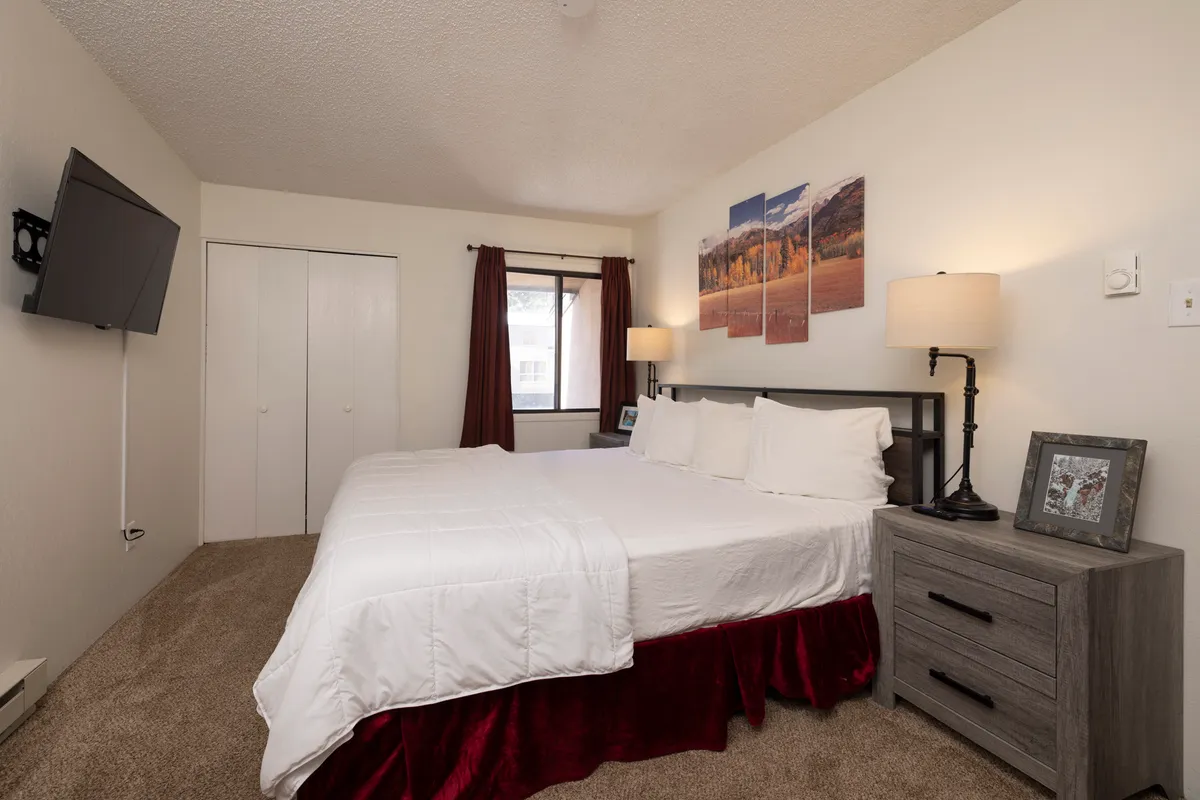 This image depicts a serene bedroom interior featuring a neatly made bed, framed artwork above the headboard, a nightstand with a lamp, and a window allowing natural light to filter in. The overall layout and calming decor convey a restful, inviting atmosphere perfect for relaxation.