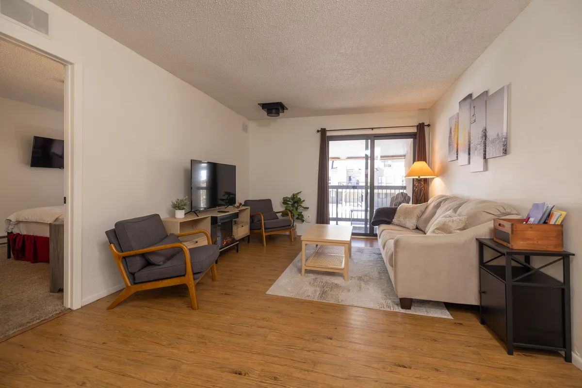 This image features a plush sofa facing a coffee table and a TV, with a balcony door letting in natural light. Artwork adorns the wall, and the space feels inviting and designed for relaxation or social interaction.
