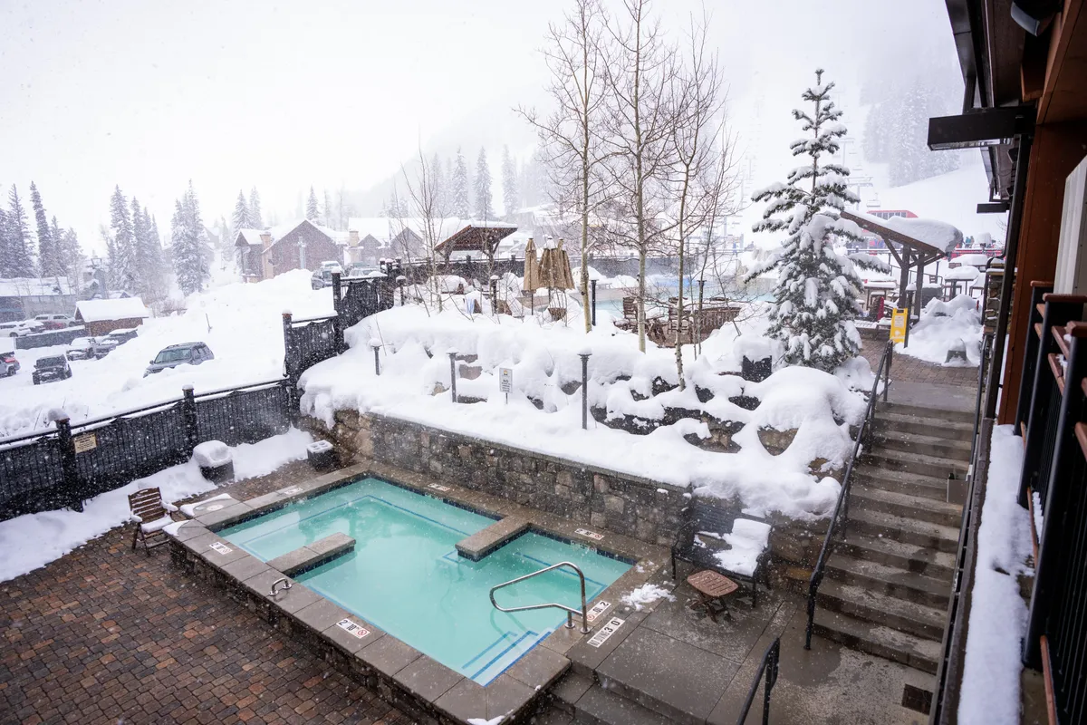 An exterior winter scene featuring the hot tub set against a snow-covered landscape, with trees and a stone pathway leading to the area. The image evokes tranquility and invites viewers to imagine relaxing in warm waters while surrounded by serene snowy views.