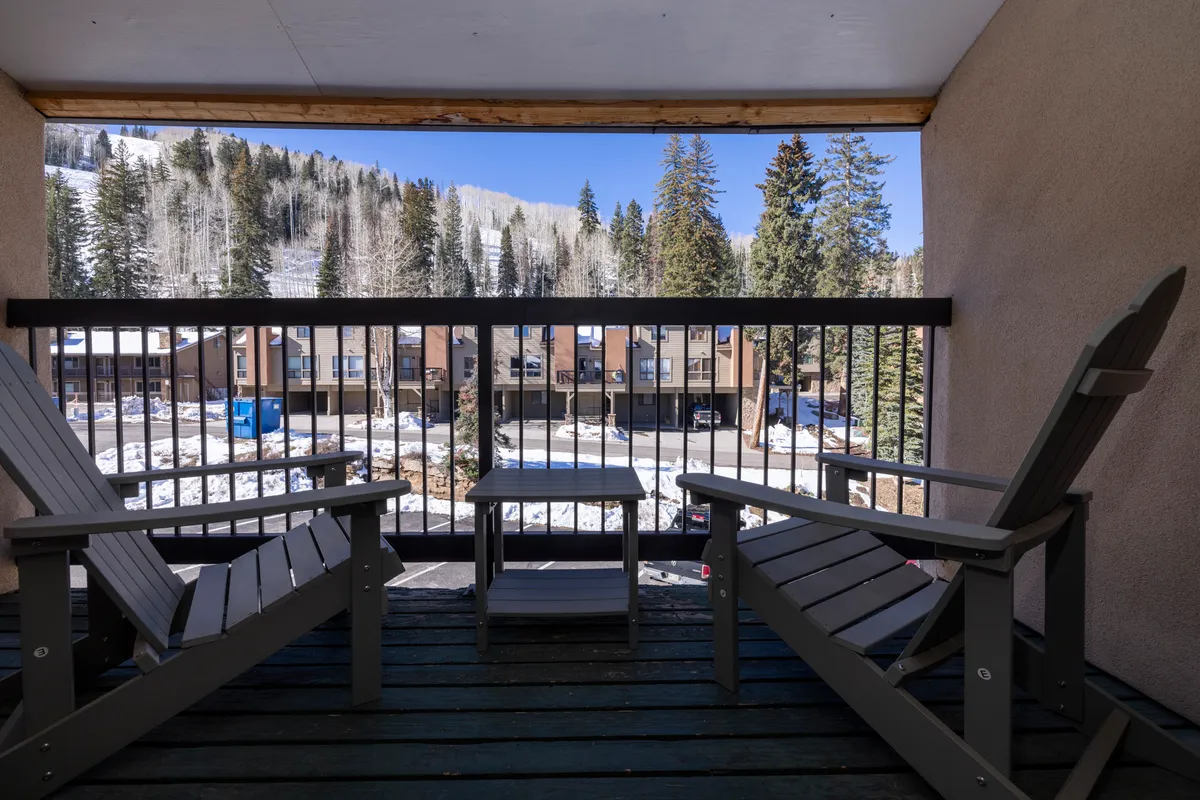 A pair of wooden chairs and a small table on a balcony overlook snowy mountains and nearby buildings. The vantage point invites viewers to imagine relaxing after a day on the slopes, with skiers visible in the distance during winter and expansive views year-round.