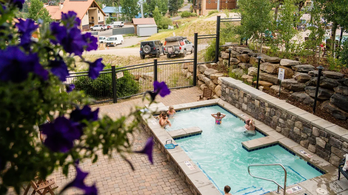 This exterior image captures both the swimming pool and hot tub at Purgatory Lodge, surrounded by flowering plants and a stone wall. The setting exudes relaxation and leisure, with people enjoying a sunny afternoon, socializing and unwinding in the amenities.