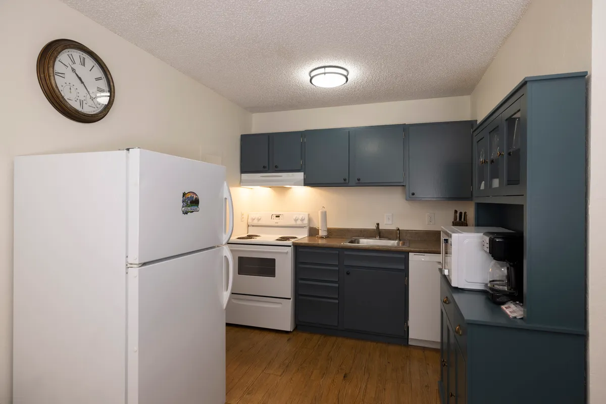 A well-lit kitchen area featuring a refrigerator, oven, sink, ample cabinetry, and a wall clock, with space for multiple people to cook or socialize. The inviting setup is perfect for both everyday meal prep and special gatherings.