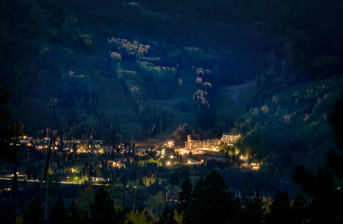 A serene nighttime view of the resort buildings softly illuminated against a backdrop of forests and distant mountains, with a clear starry sky overhead. The scene conveys the peaceful ambiance of the resort surroundings, perfect for unwinding after a day on the slopes.