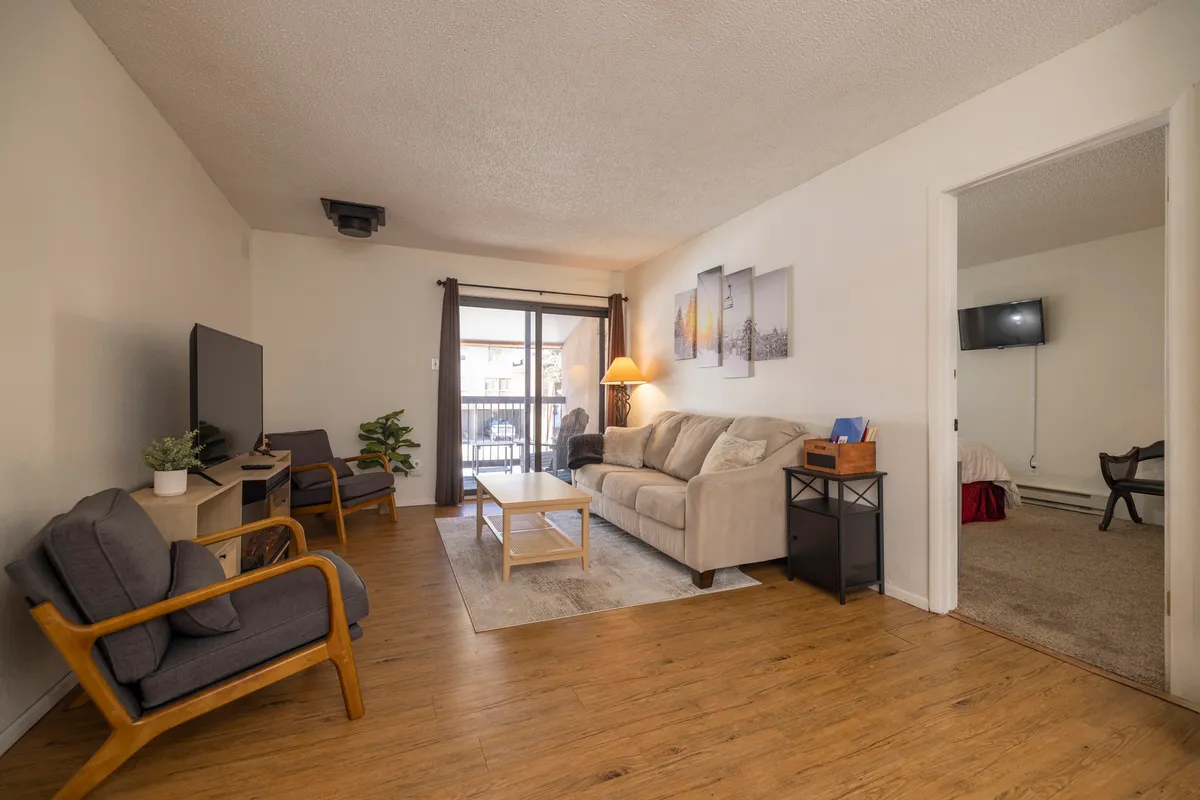 A cozy sofa faces a coffee table and television, with a balcony door providing natural light. There’s artwork on the wall and a warm, intimate ambiance, ideal for relaxation and group gatherings.