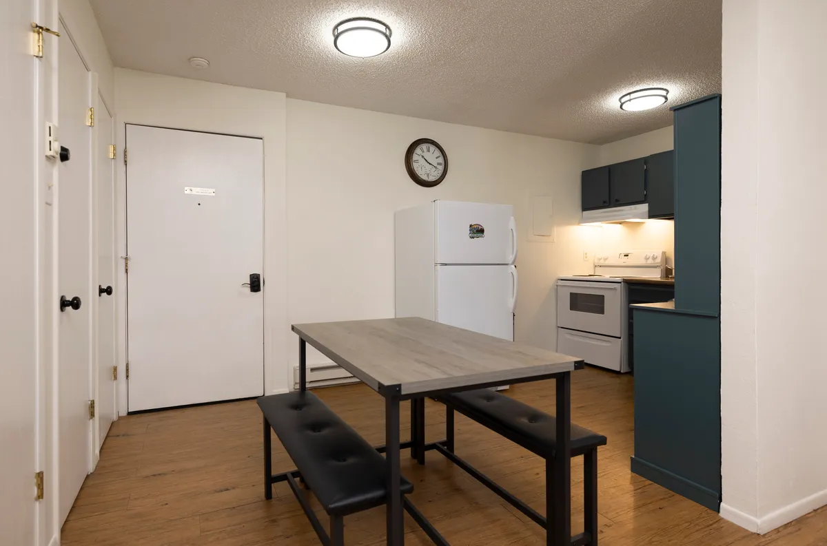 A cozy dining area with a dining table adjacent to the kitchen, a refrigerator in the background, and a wall clock. The space is clearly set for sharing meals and casual gatherings, emphasizing comfort and togetherness.