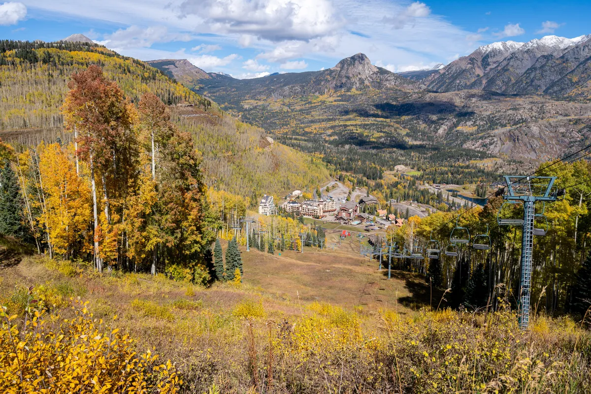 An expansive view of colorful autumn foliage set against a rugged mountain range, with resort buildings and a chairlift in clear sight. The scene suggests year-round adventure and access to both nature and local events.