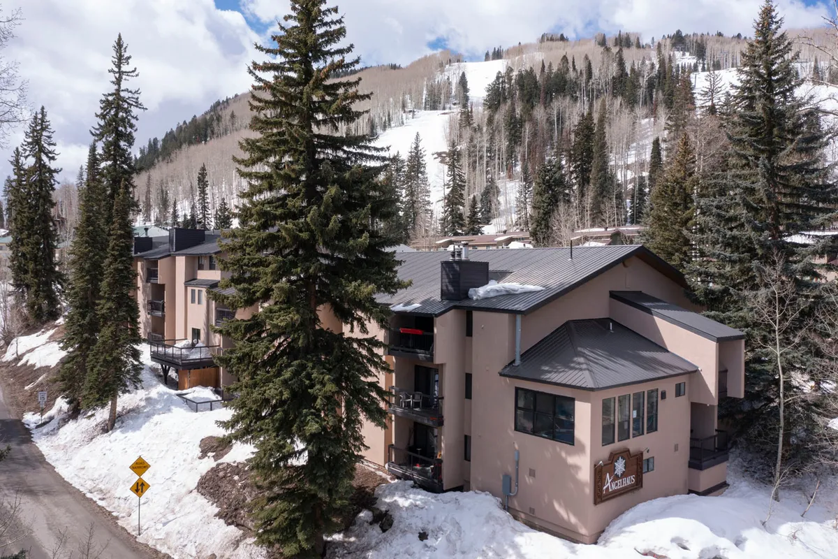 The condo building sits nestled among snowy pine trees with the majestic mountains visible behind. The scene captures both the inviting architecture and the dramatic natural setting, with subtle hints of early morning sunlight adding to the mood.